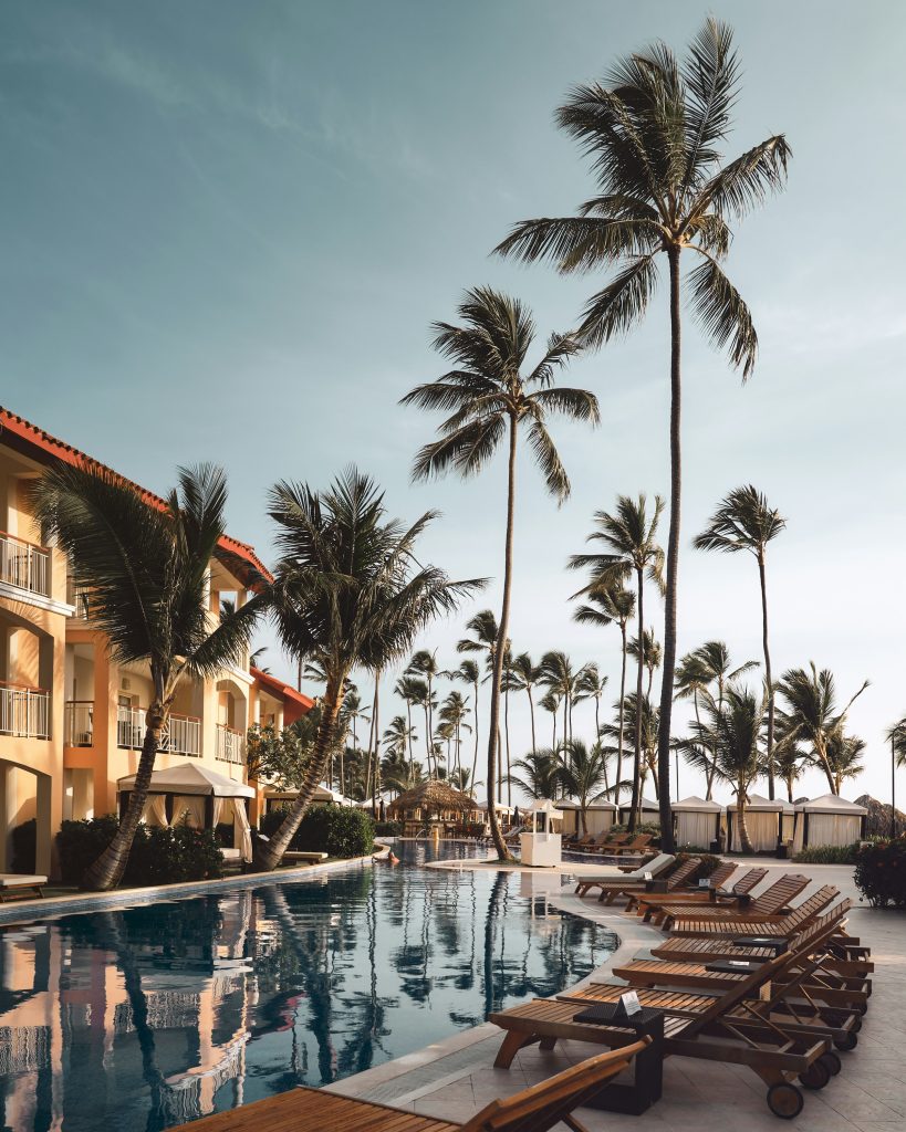 The image shows a luxurious resort pool surrounded by tall palm trees. Wooden lounge chairs are lined up along the poolside, facing the calm water, while a row of cabanas with white curtains is visible in the background. The reflection of the resort buildings, with terracotta roofs and beige exteriors, can be seen in the pool. The sky is clear with a soft hue, contributing to the tranquil, tropical atmosphere. The scene evokes a peaceful, relaxing vacation destination.