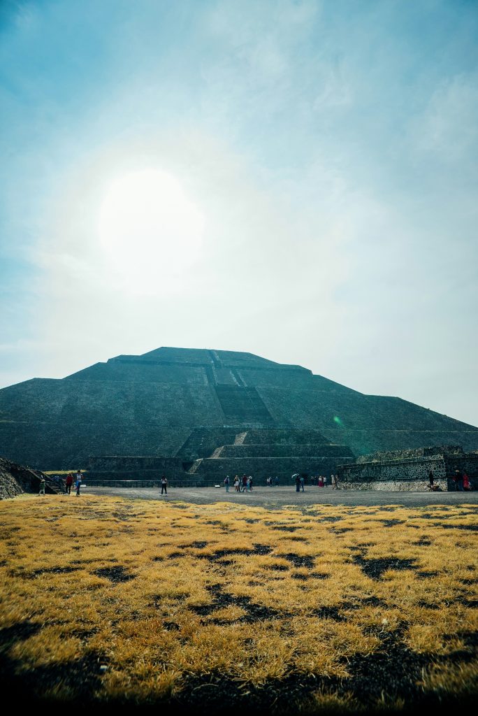 The image shows the Pyramid of the Sun at Teotihuacan, an ancient Mesoamerican archaeological site near Mexico City. The massive pyramid rises against a bright, blue sky, with the sun shining directly overhead, casting a soft light on the structure. A few visitors are scattered around the base, emphasizing the monumental scale of the pyramid. In the foreground, patches of yellow, dry grass cover the ground, contrasting with the dark stone of the pyramid. The scene captures the historical and majestic essence of this iconic landmark.