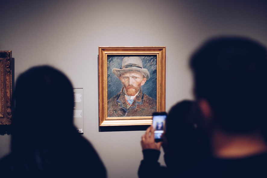 The image shows people in a museum or gallery viewing a framed portrait painting. The painting is a self-portrait of Vincent van Gogh, recognizable by his distinct style, featuring his piercing gaze, red beard, and light hat. One person in the foreground is taking a picture of the artwork with their phone, while others observe from a distance. The scene captures a moment of admiration for classic art, emphasizing the connection between modern technology and historical works in a quiet gallery setting.